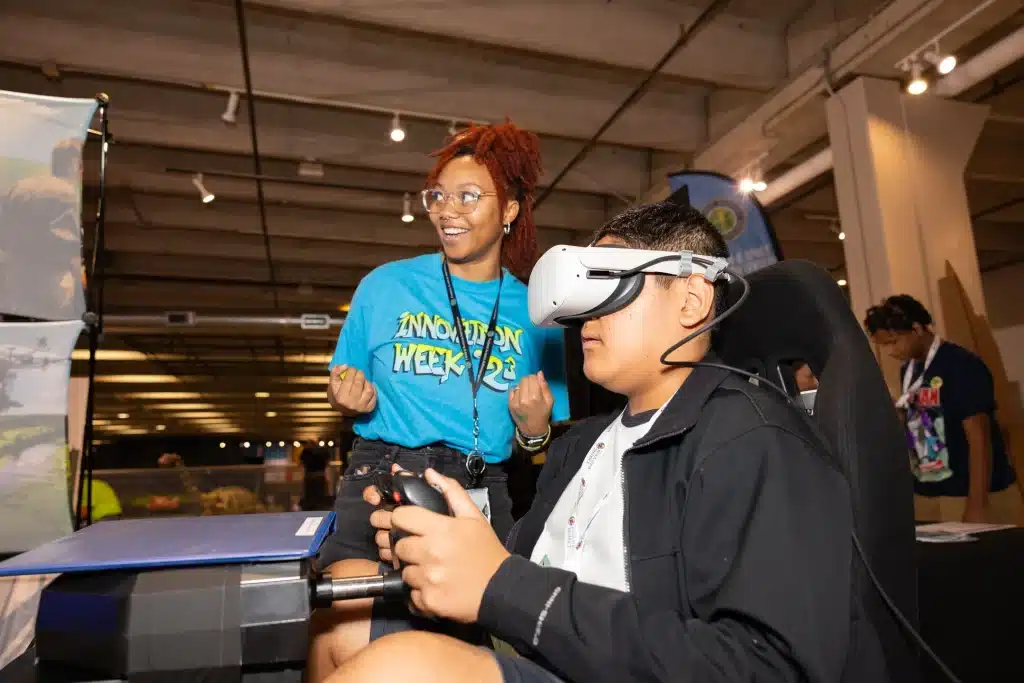 A student uses a virtual reality headset and controllers while an Innovation Week instructor looks on.
