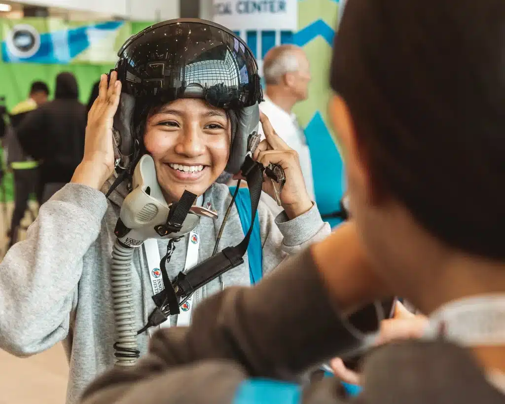 A smiling student tries on a flight helmet with help from an instructor.