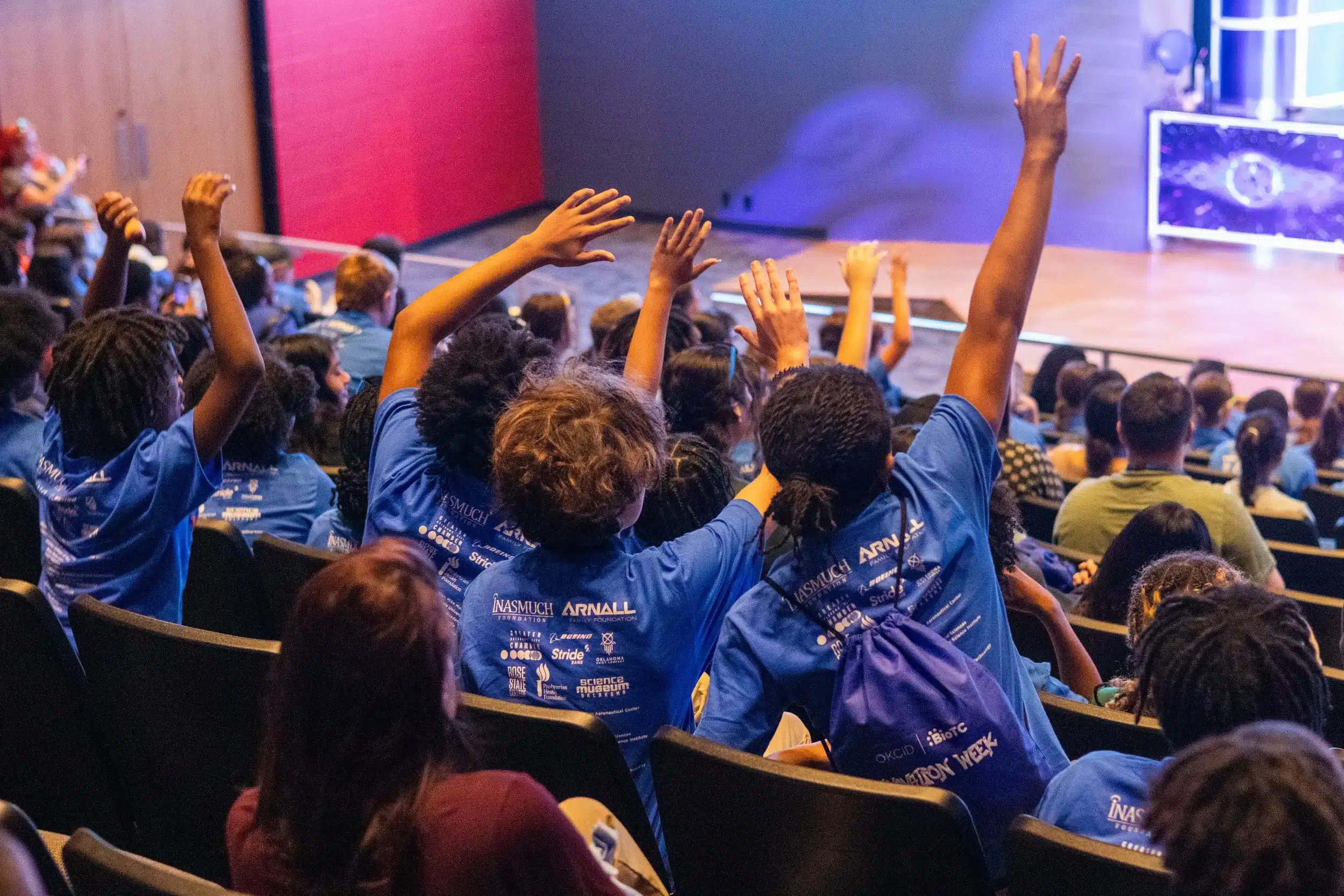 Students in Innovation Week shirts raise their hands during an auditorium presentation.