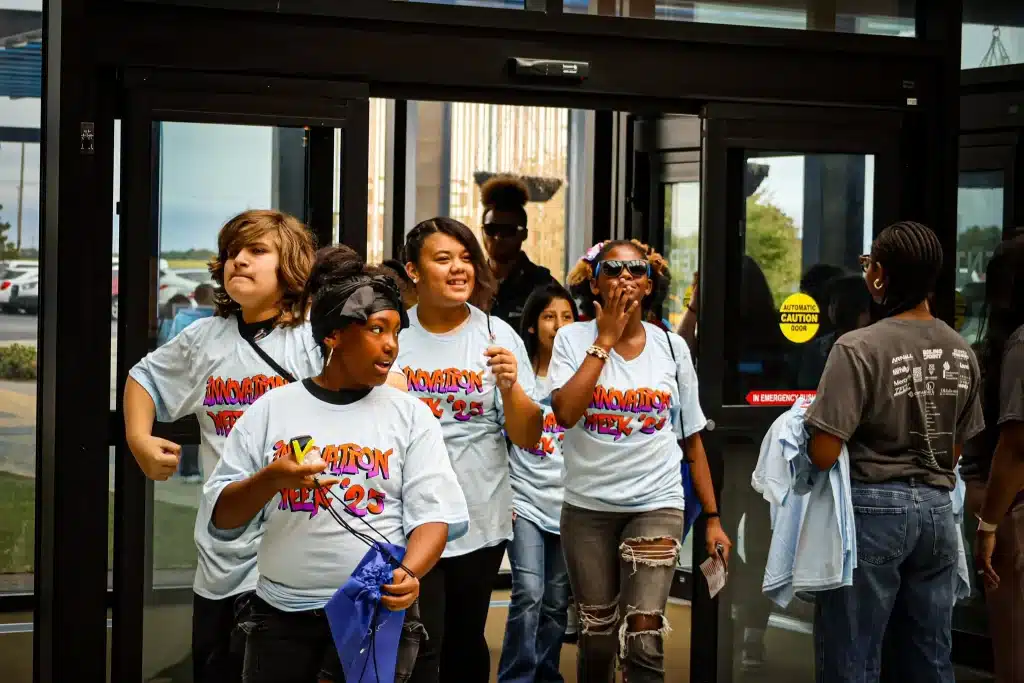 Students wearing Innovation Week 2025 shirts arrive through the building entrance.