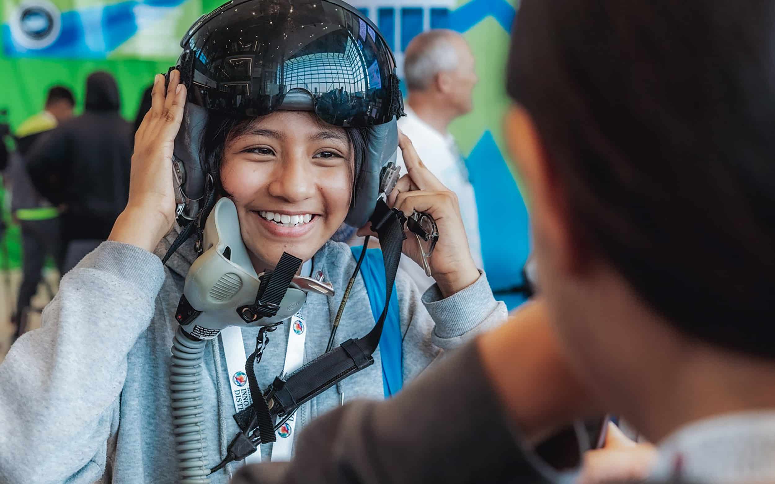 A smiling student tries on a flight helmet with help from an instructor.