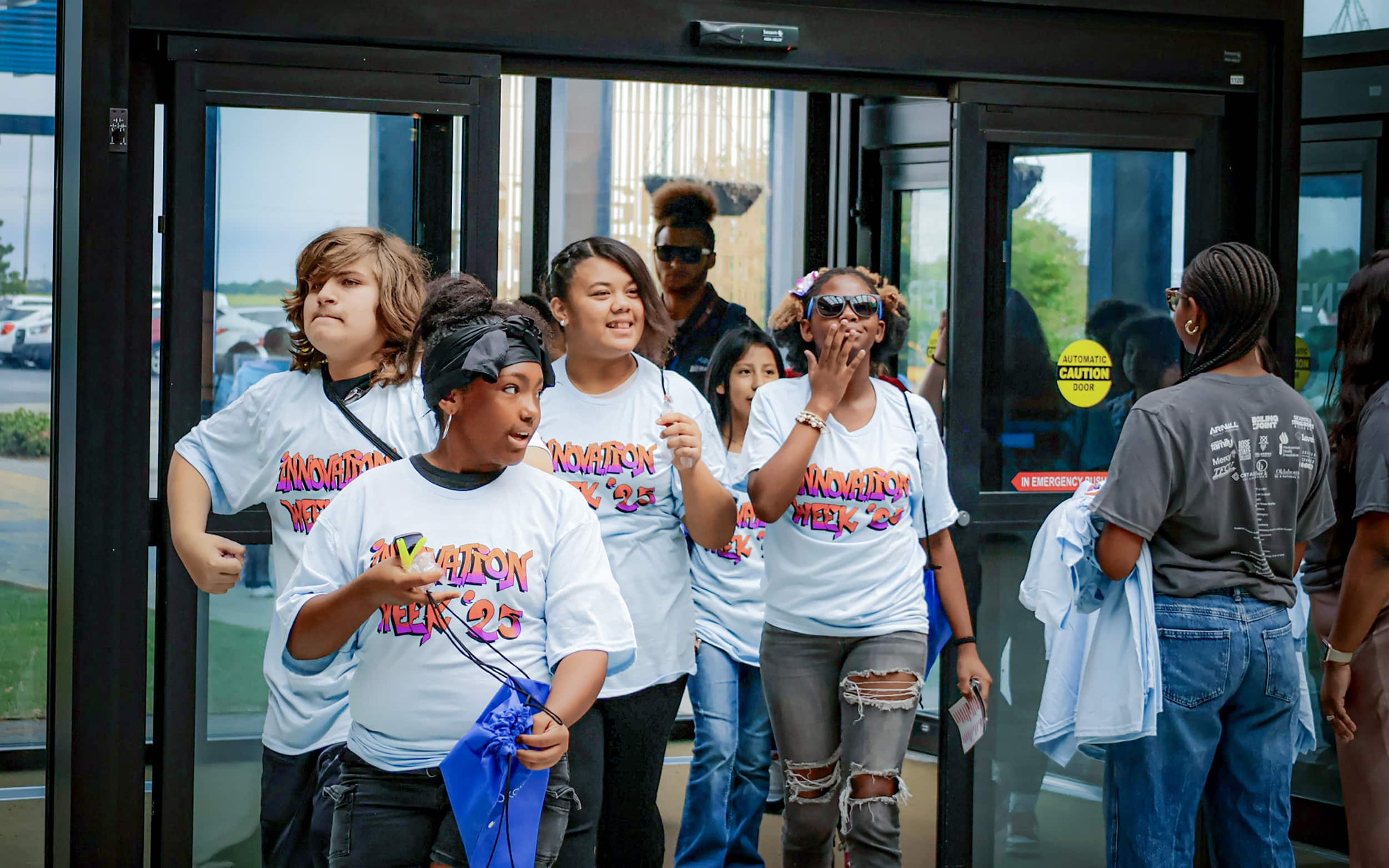 Students wearing Innovation Week 2025 shirts arrive through the building entrance.