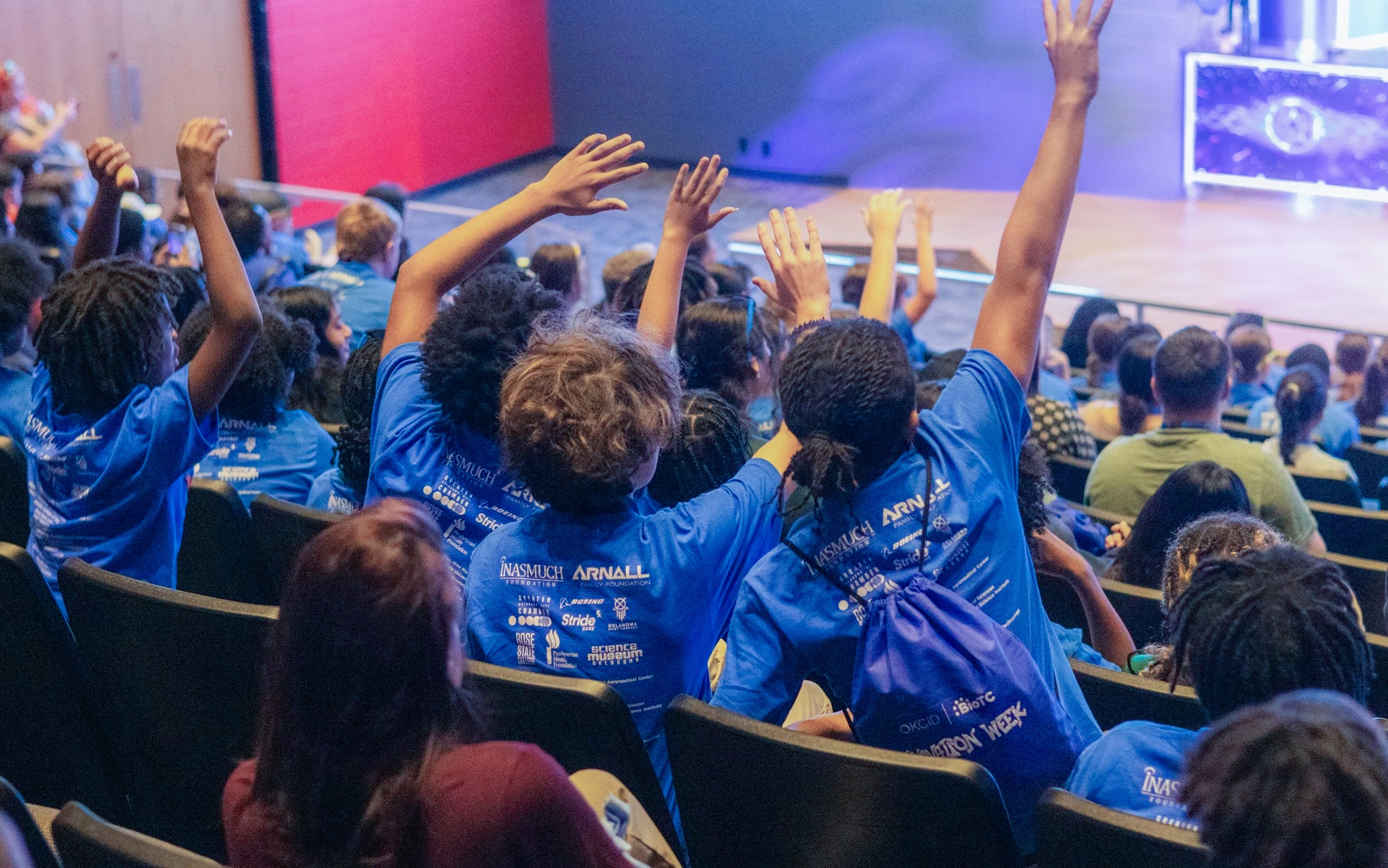 Students in Innovation Week shirts raise their hands during an auditorium presentation.