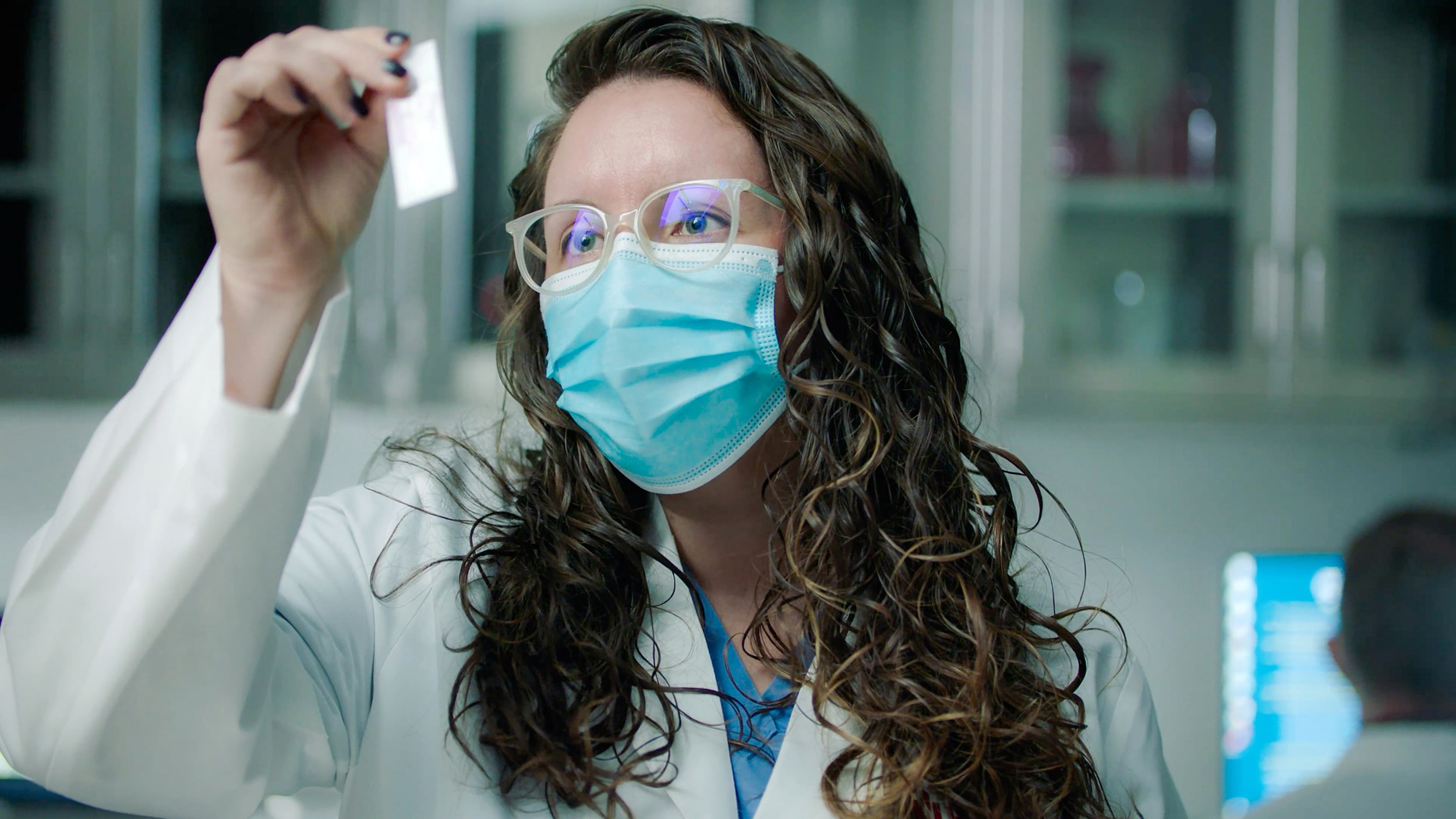 A masked laboratory researcher holds up a small sample for inspection.